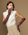 Women’s elbow-length brown leather gloves, one raised near face while wearing.