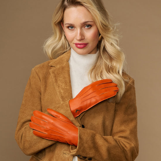 Orange leather gloves with stitched finger seams and soft cuff lining, worn with brown coat